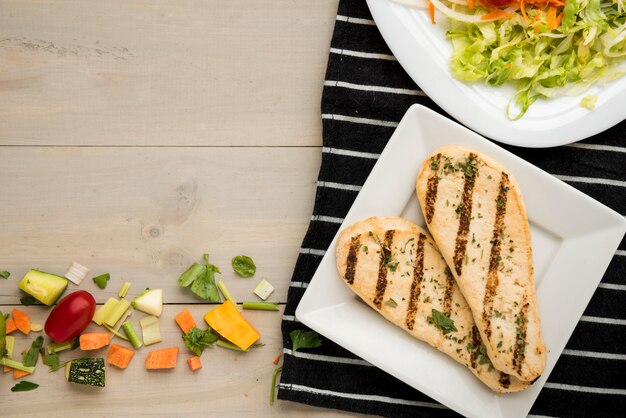 Grilled chicken fillet with salad and scattered vegetable pieces on wooden desk