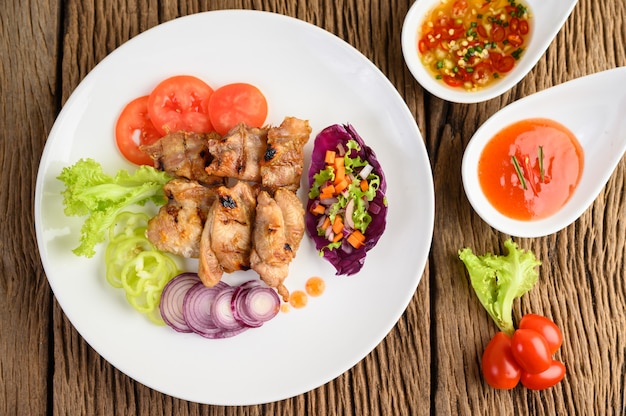 Grilled chicken on a white plate with a salad, tomatoes, chilies cut into pieces and sauce on wooden table.