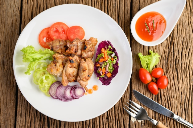 Grilled chicken on a white plate with a salad, tomatoes, chilies cut into pieces and sauce on wooden table.