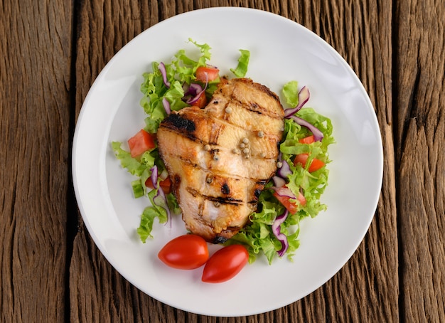 Grilled chicken on a white plate with a salad, tomatoes, chilies cut into pieces on wooden table.