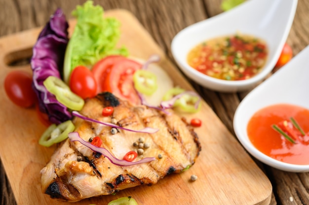 Grilled chicken on a Wood cutting board with salad, tomatoes, chilies cut into pieces, and sauce.