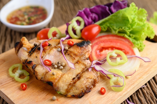 Жареная курица на деревянной разделочной доске с салатом, помидорами, чили, нарезанными на кусочки, и соусом.