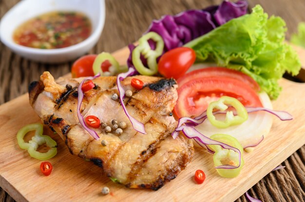 Grilled chicken on a Wood cutting board with salad, tomatoes, chilies cut into pieces, and sauce.