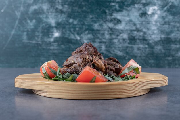 Grilled kebab and tomato slices on wooden plate.