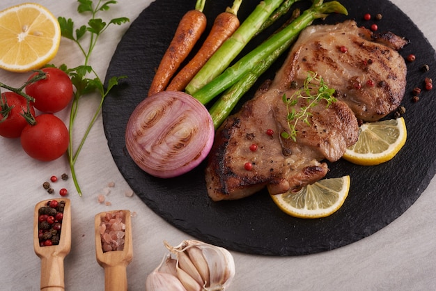 Grilled pork steak from a summer BBQ served with Vegetables, asparagus, baby carrots, fresh tomatoes and  spices. Grilled steak on  black slate on stone surface . top view.