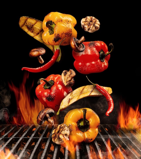 Grilled red and yellow bell pepper, zucchini, champignons, chilli and halves of garlic are falling down on black background. Bbq grill, flaming fire, charcoal and smoke. Cooking. Close up, copy space