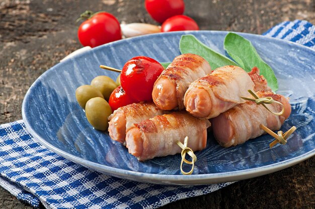 Grilled sausages wrapped in strips of bacon with tomatoes and sage leaves