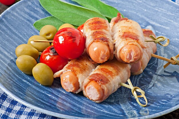 Grilled sausages wrapped in strips of bacon with tomatoes and sage leaves