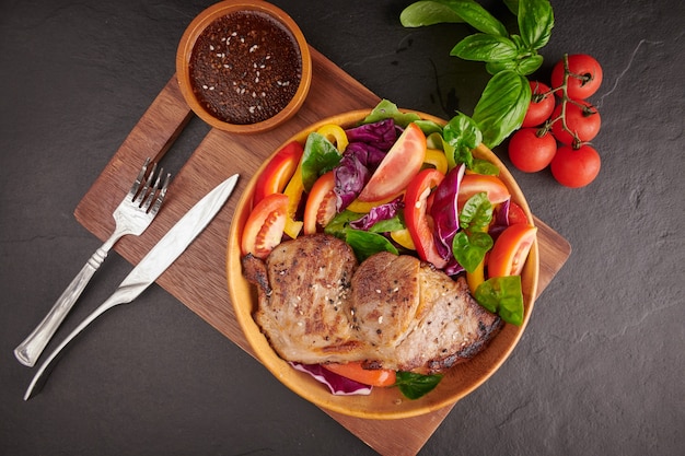 Grilled steak with fresh vegetable, sweet pepper, tomatoes, red onion, pink pepper and spices. Home made tasty food. Concept for tasty and healthy meal. Black stone surface. Pork steak with salad