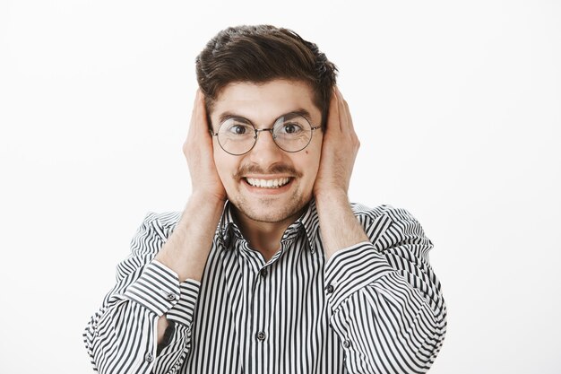 grinning excited attractive male in glasses, covering ears with palms and smiling broadly, waiting for loud noise of firework or bang, pleased and happy during party over gray wall