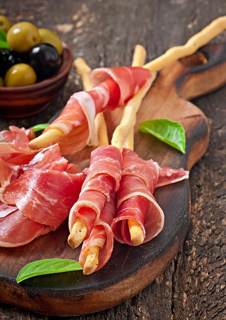 Grissini bread sticks with ham, olives, basil on old wood