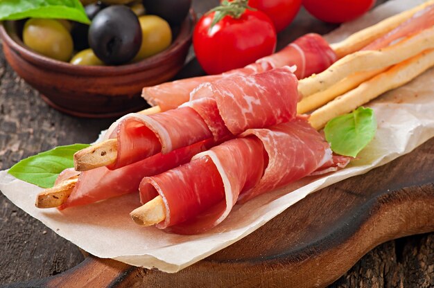 Grissini bread sticks with ham, olives, basil on old wooden