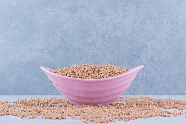 Groats spilled all over, with a full, pink bowl of buckwheat sitting in the middle on marble surface