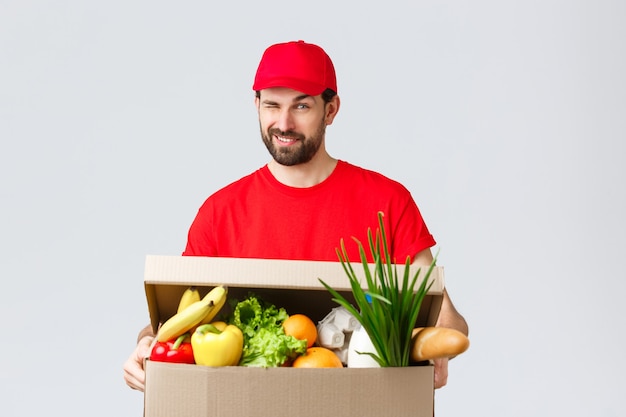 Free photo groceries and packages delivery, covid-19, quarantine and shopping concept. handsome smiling courier in red uniform, give cheeky wink as delivering food box, online order to client house.