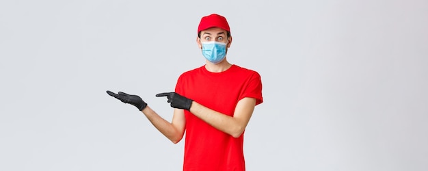 Groceries and packages delivery, covid-19, quarantine and shopping concept. Surprised and excited courier in red uniform, face mask and gloves introduce good new bonus offer, promo, showing banner.
