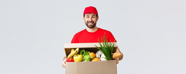 식료품 및 패키지 배달 covid19 검역 및 쇼핑 개념 빨간색 유니폼을 입은 웃는 배달원은 사람들이 물건을 옮기는 데 도움이 되는 음식 상자 소포를 들고 고객 주문을 가져옵니다.