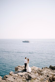 신랑과 신부는 바다를 항해하는 배를 배경으로 바위 해안에 서 있다