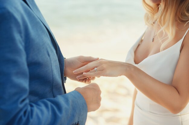 Groom in blue suit puts wedding ring on bride's hand standing on