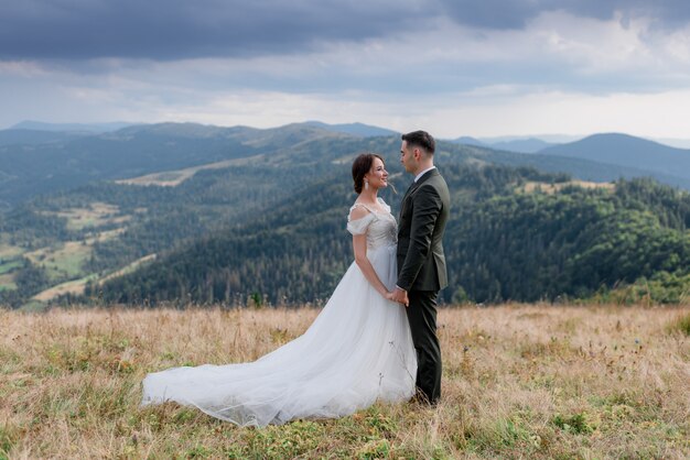 Groom and bride are standing in front of each other on the top of  a  hill in the summer mountains