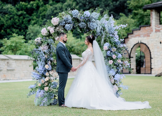 Groom and bride are standing together in front of the decorated archway with blue hydrangea, holding hands, wedding ceremony, wedding vows