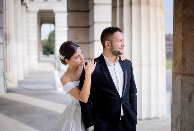 Groom and bride is standing near the huge column on the warm sunny day