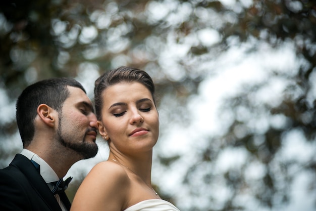 Free photo groom gently embraces the bride with a bouquet from behind in a park