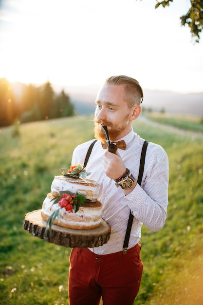 Groom holds a wooden tray with wedding cake and a pipe