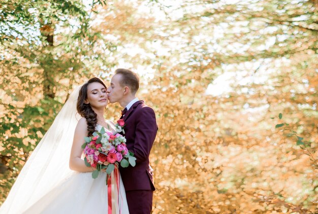 Groom is kissing bride on the cheek surrounded with yellow autumn trees