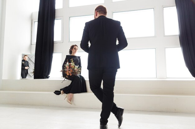 Groom's reflection in mirror. Bride sitting on stairs in white studio near window