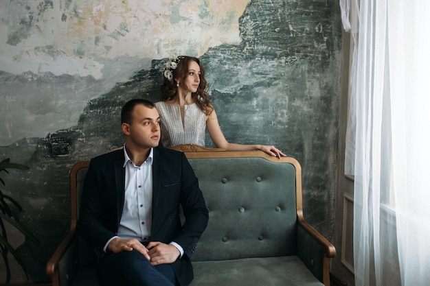 The groom sits on the sofa and bride stands near her husband