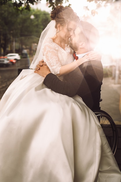 Жених на инвалидной коляске держит невесту на коленях, сидя на улице в парке