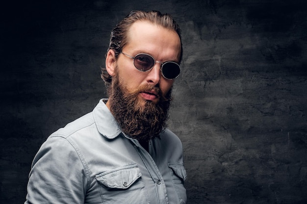 Uomo barbuto curato in camicia leggera sta posando in uno studio fotografico scuro.