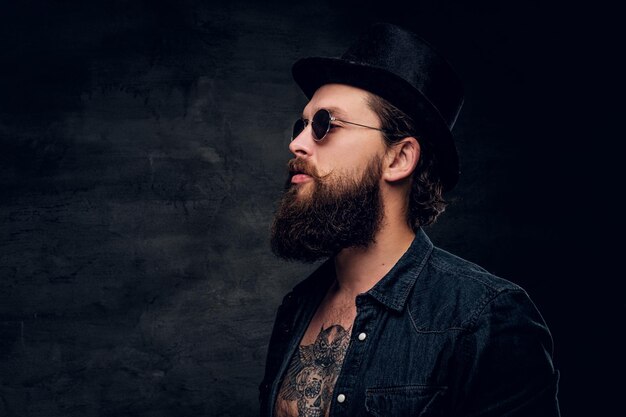 groomed bearded man with tattooed chest is posing for photographer at dark photo studio.