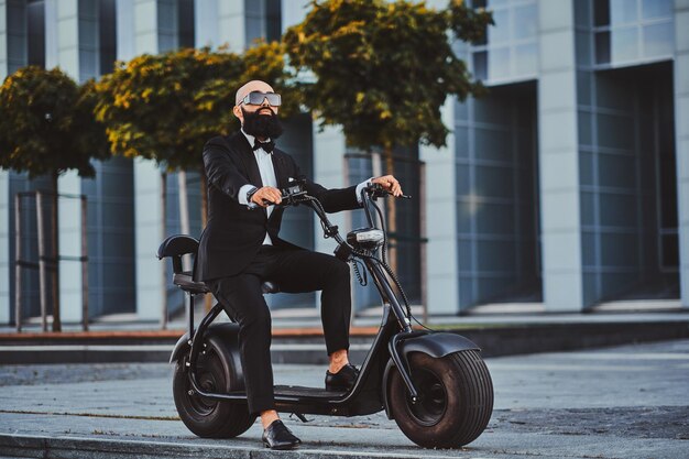 Groomed modern businessman in sunglasses is driving electro scooter to his office.