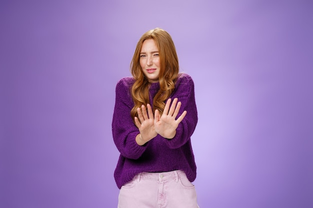 Gross take it away. Portrait of disgusted redhead woman step back and waving palms over body in refusal and no gesture grimacing and squinting from bad smell or aversion over purple wall.