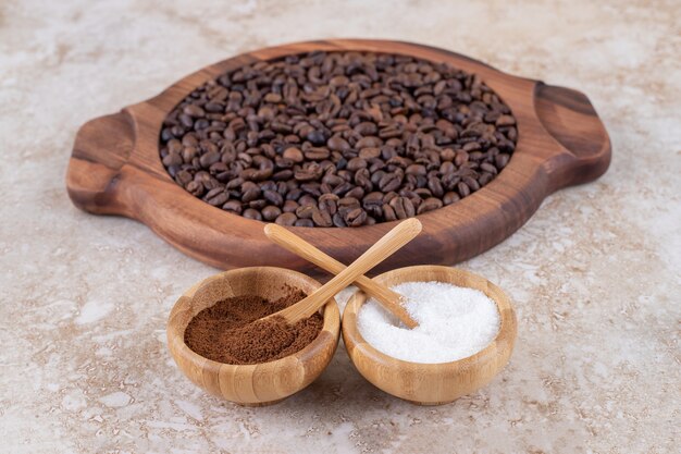 Ground coffee and a sugar bowls next to coffee beans piled on a wooden tray 