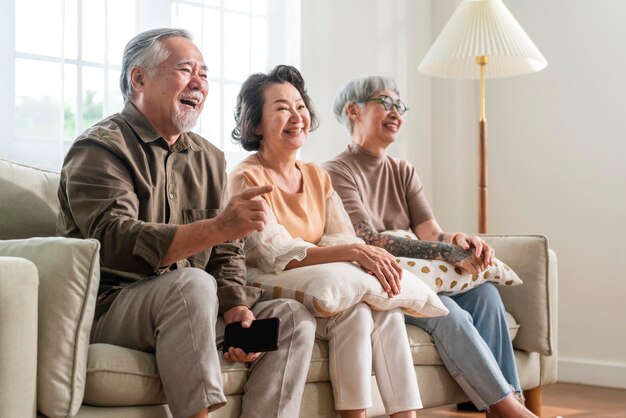 Группа азиатских пожилых мужчин и женщин, проводящих выходные вместе, сидят на диване и смотрят телевизионную комедийную программу с радостным смехом, улыбающимся выражением счастья, пожилые люди в доме престарелых