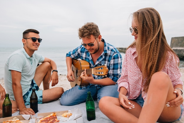 Group of attractive friends having a picnic, playing guitar on the beach, while eating pizza