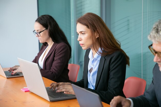 Group of business colleagues sitting in line and using computers in office. Business professionals sitting at one table and typing on laptop keyboards. Medium shot. Communication or wireless technolog