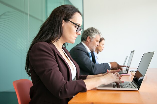 並んで座って、オフィスでコンピューターを使用しているビジネス人々のグループ。ノートパソコンのキーボードで入力するさまざまな年齢の従業員。