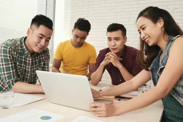 Group of casually dressed young Asian people standing around table and looking at laptop screen 