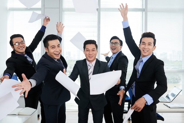 Group of cheerful Asian businessmen in suits throwing documents up in air in office