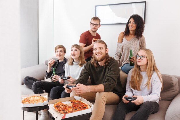 Gruppo di amici allegri che trascorrono del tempo insieme mentre giocano ai videogiochi, mangiano pizza e bevono birra a casa