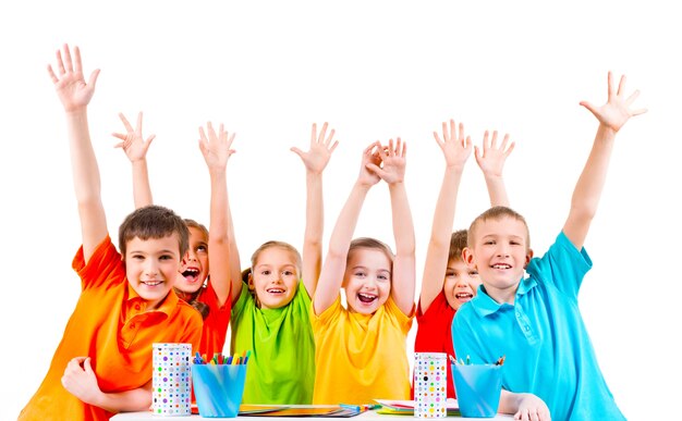 Group of children in colored t-shirts sitting at a table with raised hands.