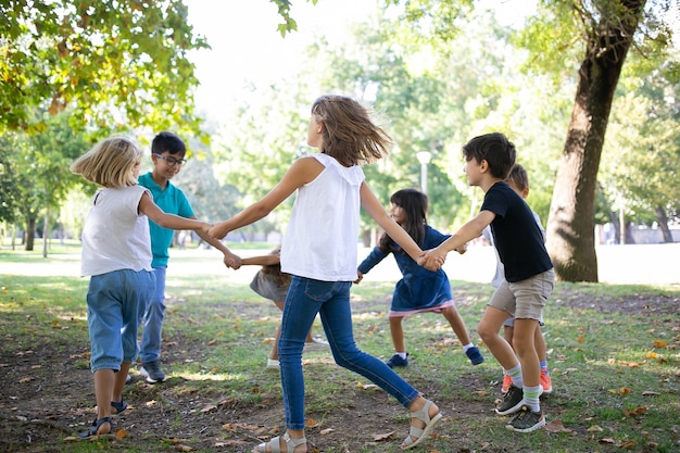 Группа детей, взявшись за руки и танцующих, наслаждаясь мероприятиями на свежем воздухе и весело проводящими время в парке. Детская вечеринка или концепция дружбы