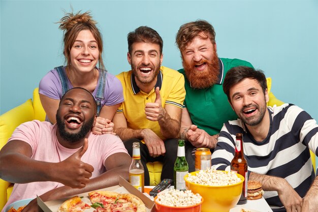 Group of diverse friends cheer as favorite team wins, show thumb up gesture, eat tasty pizza and popcorn, smile broadly, drink beer, isolated over blue wall. People, entertainment, fun concept