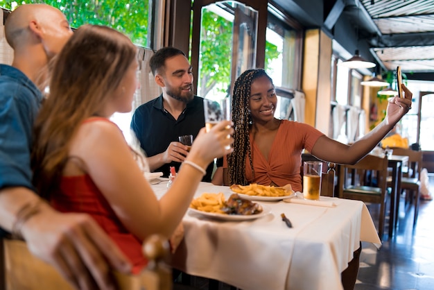 バーでビールを飲みながら携帯電話で自分撮りをしている多様な友人のグループ。友達のコンセプト。