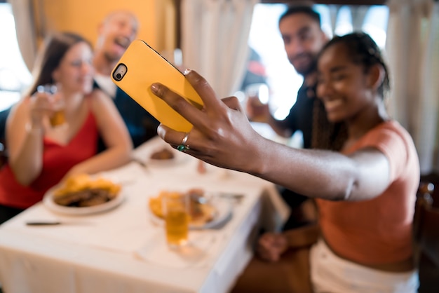 レストランで一緒に食事を楽しみながら携帯電話で自分撮りをしている多様な友人のグループ。友達のコンセプト。