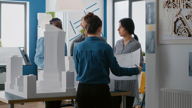 Группа разнообразных людей, работающих над дизайном модели здания с ноутбуком и чертежами. Многонациональные коллеги используют технологии и строительный план для планирования архитектурной стратегии.