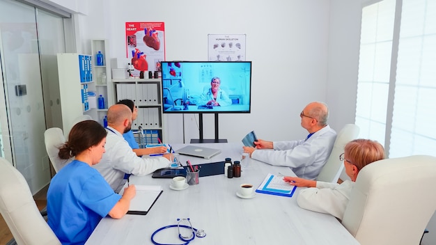 Group of doctors discussing with expert medic during video conference from hospital office. Medicine staff using internet during online meeting with expert doctor for expertise, nurse taking notes.